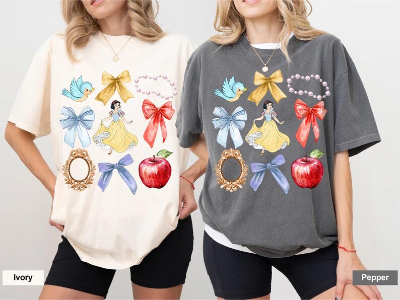 K&ouml;nnte beinhalten: Zwei T-Shirts mit einem Schneewittchen-Motiv. Die Shirts zeigen verschiedene Bilder, darunter Schneewittchen, V&ouml;gel, Schleifen, &Auml;pfel und Spiegel. Die Shirts sind in Elfenbein und Pfeffer erh&auml;ltlich.