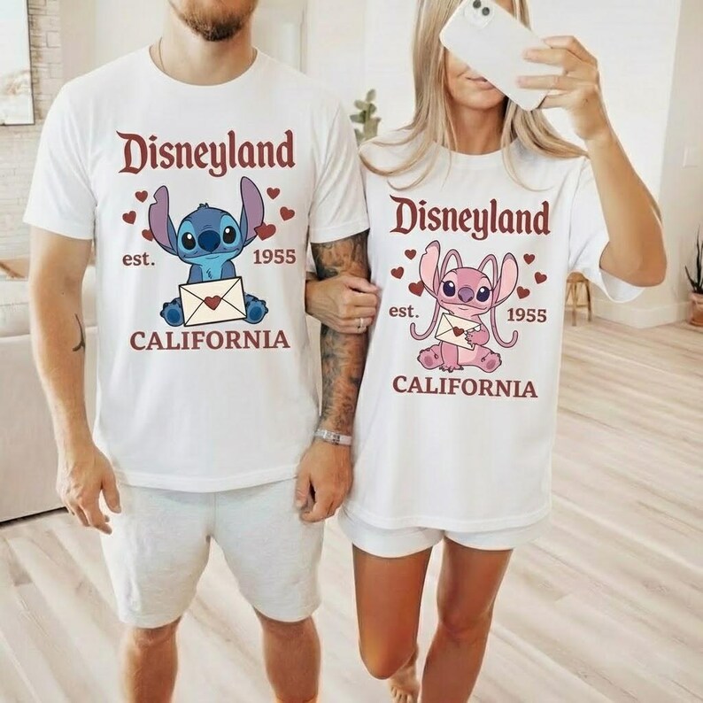 Op de afbeelding: Witte T-shirts met Disneyland-afbeeldingen. E&eacute;n shirt heeft een blauw stripfiguur met een envelop, de andere een roze figuur. Beide shirts hebben de tekst "Disneyland est. 1955 CALIFORNIA" in bordeaux.