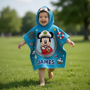 Op de afbeelding: Een lichtblauwe poncho met capuchon met een lachend kind. De poncho heeft een Mickey Mouse-ontwerp in een patrijspoort, nautische elementen en de naam "JAMES". De capuchon is versierd met een Mickey Mouse-gezicht.
