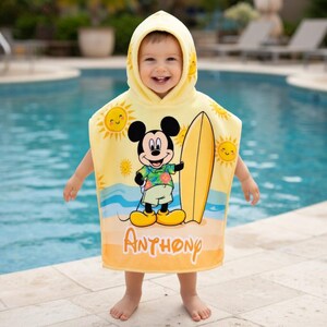 Puede incluir: Una toalla de playa con capucha amarilla con Mickey Mouse sosteniendo una tabla de surf. La toalla tiene una escena de playa soleada con el nombre "Anthony" impreso. La toalla est&aacute; dise&ntilde;ada para un ni&ntilde;o.