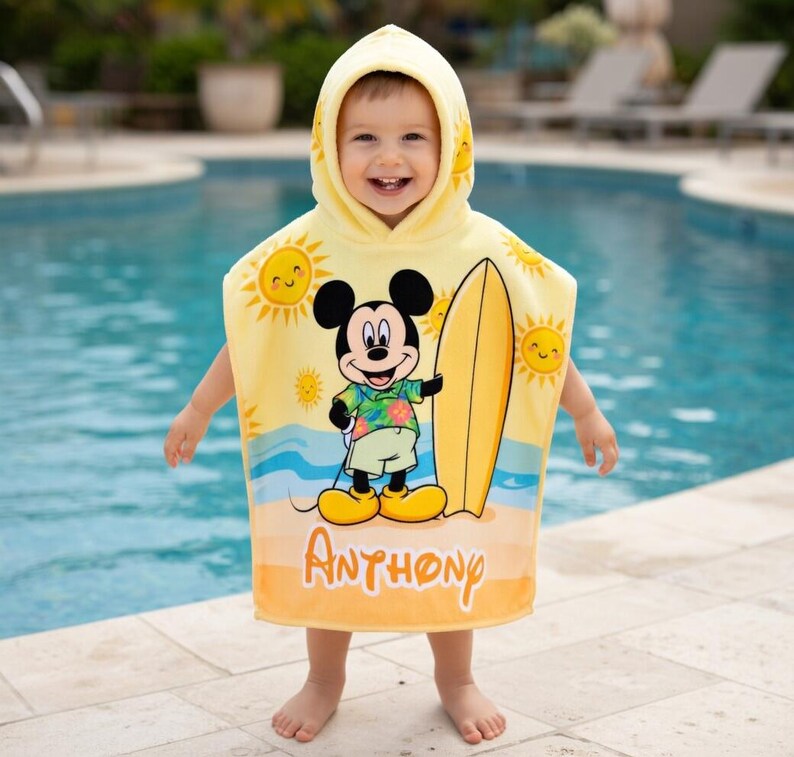 Puede incluir: Una toalla de playa con capucha amarilla con Mickey Mouse sosteniendo una tabla de surf. La toalla tiene una escena de playa soleada con el nombre "Anthony" impreso. La toalla est&aacute; dise&ntilde;ada para un ni&ntilde;o.