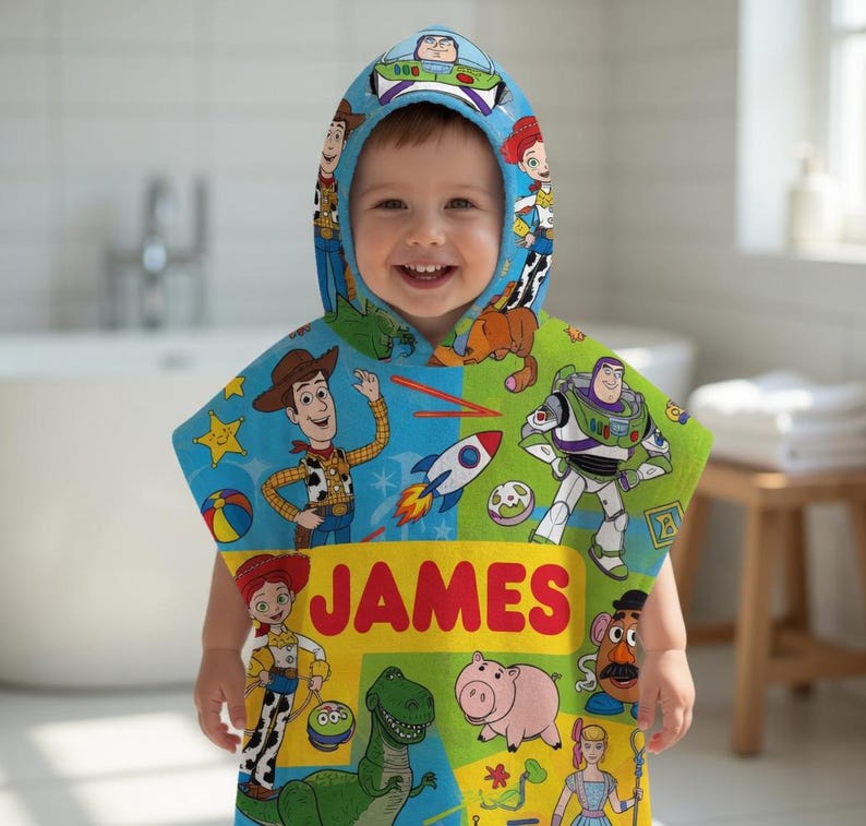 Puede incluir: Una toalla con capucha y colorida con personajes de la pel&iacute;cula Toy Story. La toalla es azul y verde con im&aacute;genes de Woody, Buzz Lightyear y otros personajes. El nombre "JAMES" est&aacute; impreso en la parte delantera.