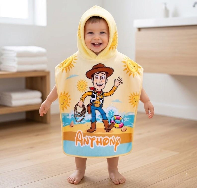 Puede incluir: Una toalla de playa con capucha amarilla con una imagen de dibujos animados de Woody de Toy Story, con gr&aacute;ficos de sol y playa. La toalla tiene el nombre "Anthony" impreso en la parte inferior. El ni&ntilde;o lleva la toalla.