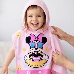 May include: A pink hooded towel featuring a Minnie Mouse graphic wearing sunglasses and a floatie. The towel has a hood and is being held by a person. The Minnie Mouse graphic is in the center of the towel.