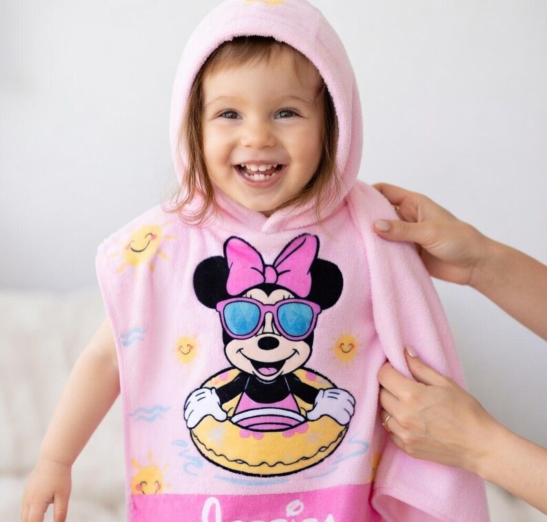 May include: A pink hooded towel featuring a Minnie Mouse graphic wearing sunglasses and a floatie. The towel has a hood and is being held by a person. The Minnie Mouse graphic is in the center of the towel.