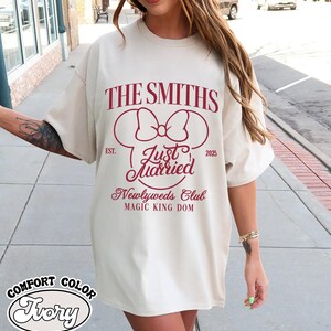 May include: Ivory-colored t-shirt with the text "THE SMITHS" and "Just Married" in burgundy. The design includes a bow and Mickey Mouse ears, with "EST. 2025" and "Newlyweds Club MAGIC KING DOM" below. The shirt is labeled "COMFORT COLOR Ivory".