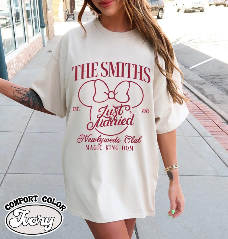 May include: Ivory-colored t-shirt with the text "THE SMITHS" and "Just Married" in burgundy. The design includes a bow and Mickey Mouse ears, with "EST. 2025" and "Newlyweds Club MAGIC KING DOM" below. The shirt is labeled "COMFORT COLOR Ivory".