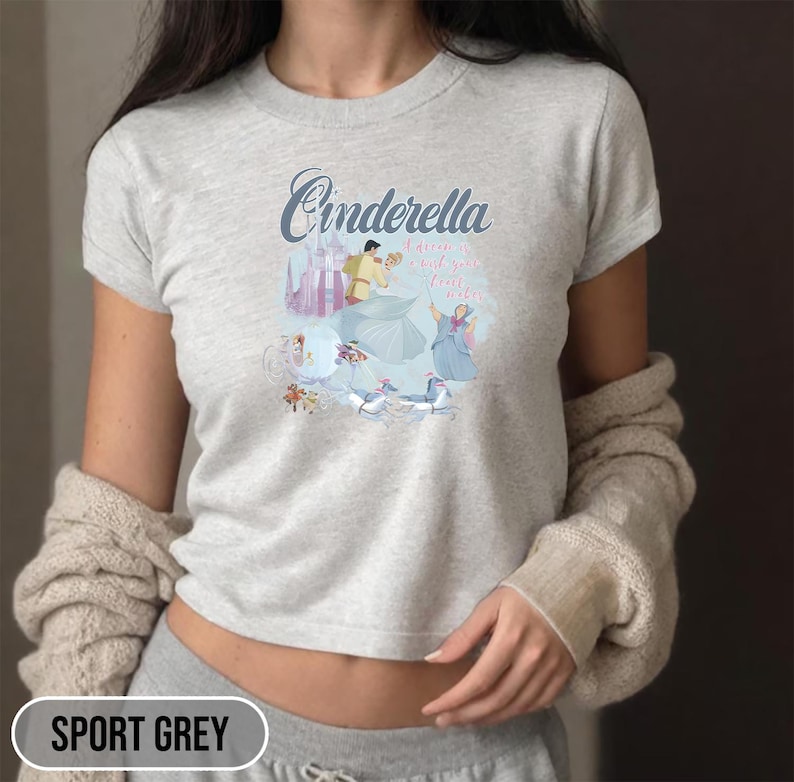 May include: A light gray t-shirt with a graphic of Cinderella and Prince Charming dancing. The text "Cinderella is a wish your heart makes" is printed on the shirt.