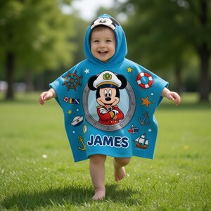 Op de afbeelding: Een lichtblauwe poncho met capuchon met een lachend Mickey Mouse kapitein-ontwerp. De poncho heeft nautische versieringen, waaronder een scheepsstuur, anker en een reddingsboei. De naam "JAMES" staat op de voorkant.