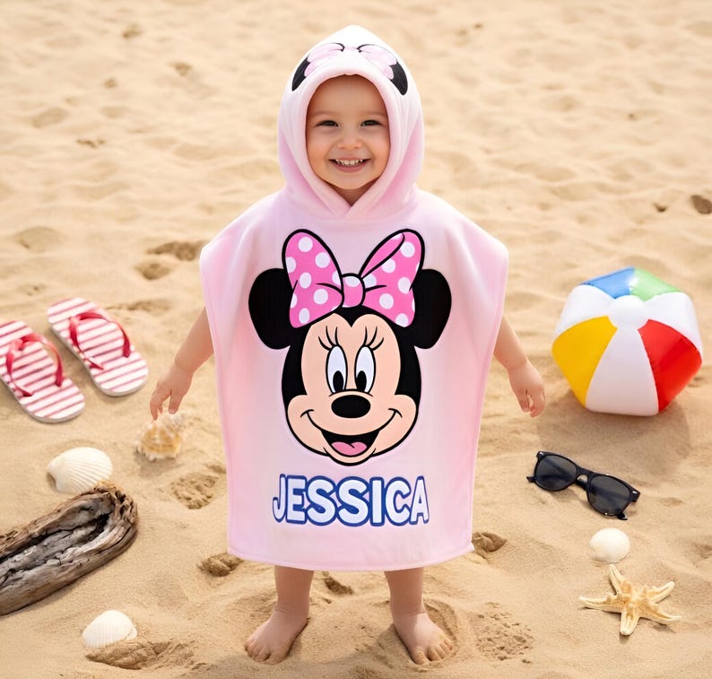 May include: A child wearing a pink hooded towel featuring Minnie Mouse with a polka dot bow and the name "JESSICA". The towel is on a sandy beach, with flip-flops, a beach ball, and sunglasses nearby.