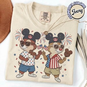 May include: Ivory-colored Comfort Colors t-shirt featuring a cartoon illustration of two chipmunks wearing Mickey Mouse hats and sunglasses, holding American flags and chocolate treats. The shirt has a crew neck and short sleeves. The design includes fireworks and stars.