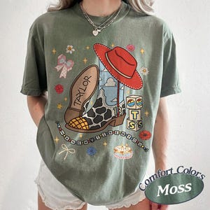 May include: An olive green t-shirt featuring a graphic print of a cow-print cowboy boot, a red hat, alphabet blocks spelling TS, a cake, and the text TAYLOR and I KNEW YOU WERE TROUBLE. The design includes small stars, flowers, and bows.