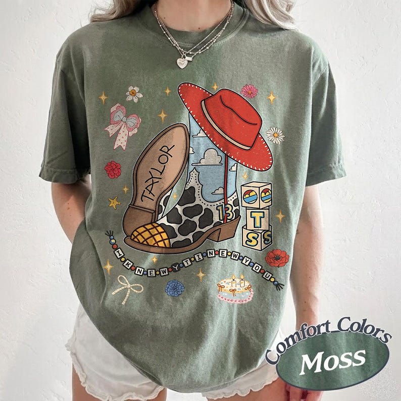 May include: An olive green t-shirt featuring a graphic print of a cow-print cowboy boot, a red hat, alphabet blocks spelling TS, a cake, and the text TAYLOR and I KNEW YOU WERE TROUBLE. The design includes small stars, flowers, and bows.