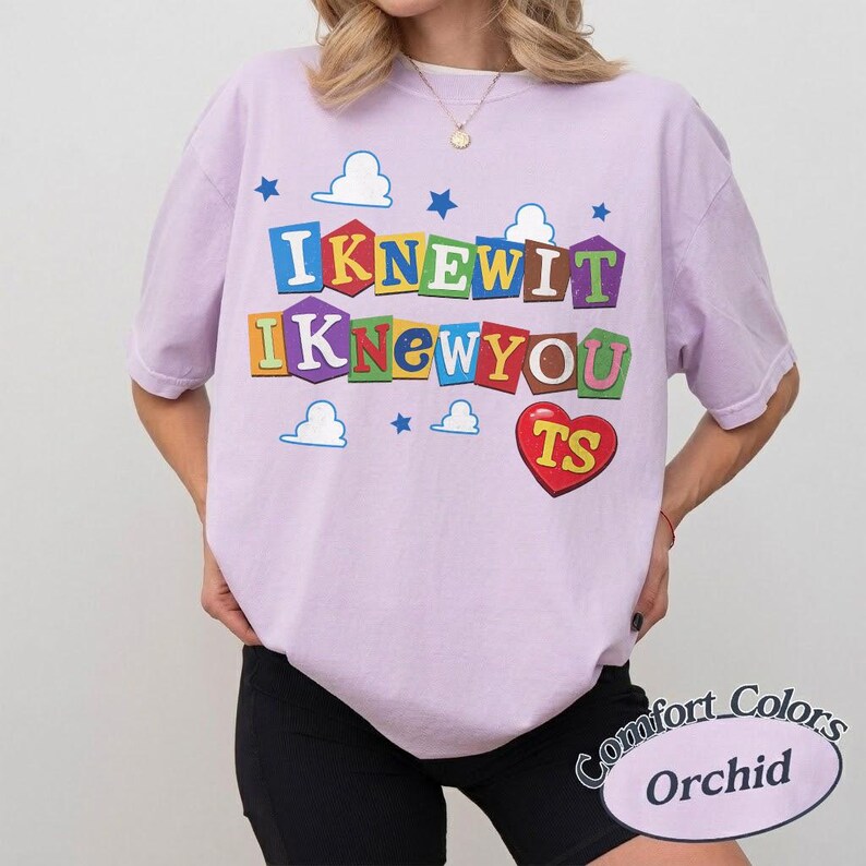 May include: A light purple short-sleeve t-shirt featuring a graphic print with the text "I KNEW IT I KNEW YOU" in colorful block letters. The design includes small blue stars and white cloud shapes, along with a red heart containing the letters "TS".