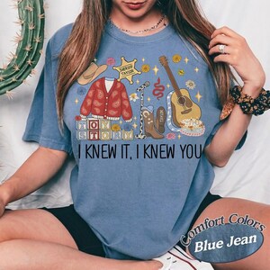 May include: A blue t-shirt featuring a graphic print with a red paisley cardigan, a sheriff badge labeled Taylor, a microphone, a brown cowboy boot, a guitar, and blocks spelling Toy Story. The text I KNEW IT, I KNEW YOU is printed below the graphic.