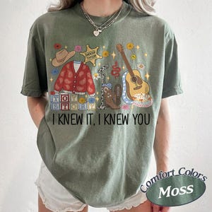 May include: A moss green short-sleeve t-shirt featuring a graphic print of a cowboy hat, a red paisley cardigan, a sheriff badge, a microphone, a brown cowboy boot, a guitar, and toy blocks. The text reads I KNEW IT, I KNEW YOU.