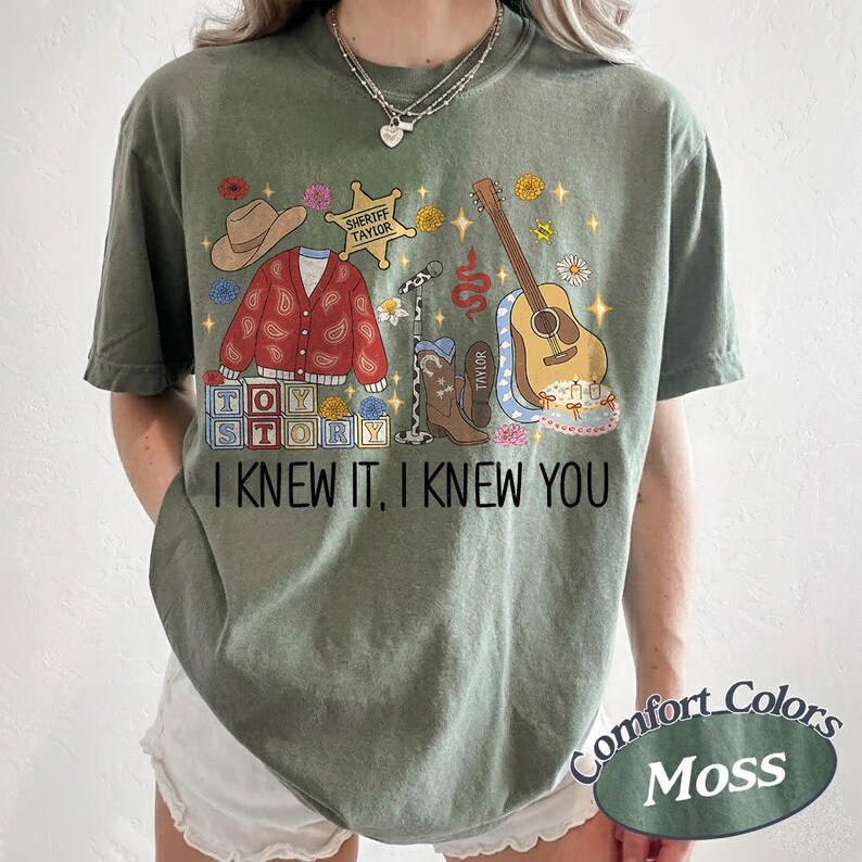 May include: A moss green short-sleeve t-shirt featuring a graphic print of a cowboy hat, a red paisley cardigan, a sheriff badge, a microphone, a brown cowboy boot, a guitar, and toy blocks. The text reads I KNEW IT, I KNEW YOU.