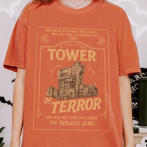 May include: Orange short-sleeved t-shirt with a vintage graphic of the Hollywood Tower of Terror. The graphic includes the text "TOWER OF TERROR" and other phrases. The shirt is a solid orange colour.