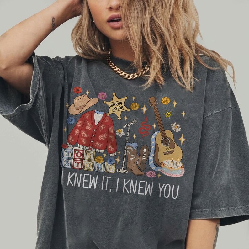May include: A charcoal gray t-shirt featuring a graphic print with a cowboy hat, a red paisley cardigan, a sheriff badge labeled "SHERIFF TAYLOR," a microphone, a guitar, cowboy boots, and blocks spelling "TOY STORY" with the text "I KNEW IT, I KNEW YOU."