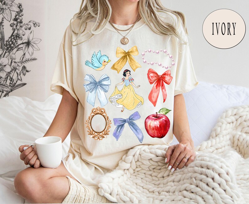 K&ouml;nnte beinhalten: Ein wei&szlig;es T-Shirt mit einem Aufdruck von Schneewittchen und verschiedenen Bildern aus dem Film Schneewittchen und die sieben Zwerge. Die Bilder beinhalten einen blauen Vogel, einen roten Apfel, einen Spiegel, eine Perlenkette und mehrere Schleifen. Das Shirt wird von einem Model getragen.