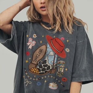 May include: A charcoal gray short-sleeve t-shirt featuring a graphic print of a cow-print cowboy boot, a red hat, blocks with the letters T, S, and the number 13, a bow, and a cake. The text TAYLOR and I KNEW YOU WERE TROUBLE are printed.