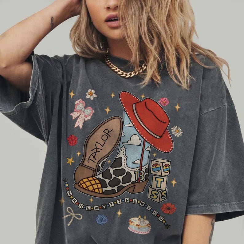 May include: A charcoal gray short-sleeve t-shirt featuring a graphic print of a cow-print cowboy boot, a red hat, blocks with the letters T, S, and the number 13, a bow, and a cake. The text TAYLOR and I KNEW YOU WERE TROUBLE are printed.