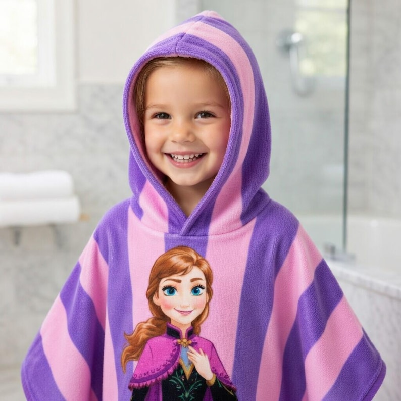 Puede incluir: Una toalla con capucha para ni&ntilde;os con rayas verticales rosas y moradas. La toalla presenta un gr&aacute;fico de un personaje de dibujos animados en la parte delantera. El ni&ntilde;o sonr&iacute;e y lleva la toalla en un ba&ntilde;o.