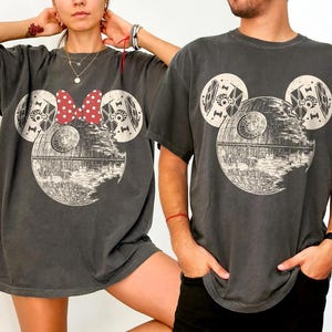 May include: Two dark gray t-shirts featuring a Death Star design in the shape of Mickey Mouse. One shirt has a red and white polka dot bow. The shirts are short-sleeved and worn by two people.