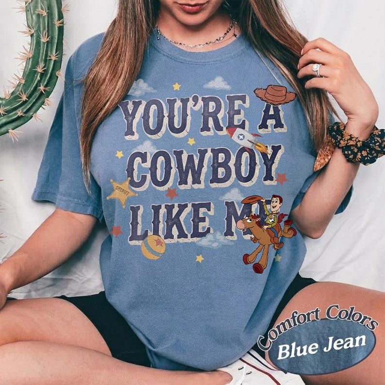 May include: A blue short-sleeve t-shirt featuring the text "YOU'RE A COWBOY LIKE ME" in a distressed font. The graphic includes a cartoon cowboy riding a horse, a rocket ship, a cowboy hat, a ball, and stars against a background of clouds.
