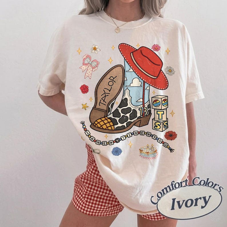 May include: An ivory t-shirt featuring a graphic print of a cow-print cowboy boot with the name Taylor, a red cowboy hat, a stack of blocks with the letters T, S, and a cake, a beaded string with the text I KNEW YOU WERE TROUBLE, and flowers.