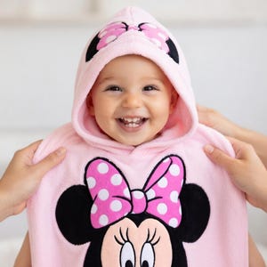 May include: A smiling baby wearing a pink hooded towel with a Minnie Mouse design. The towel features a large polka-dot bow and Minnie's face. The hood has a matching bow design. The baby is being held by two hands.