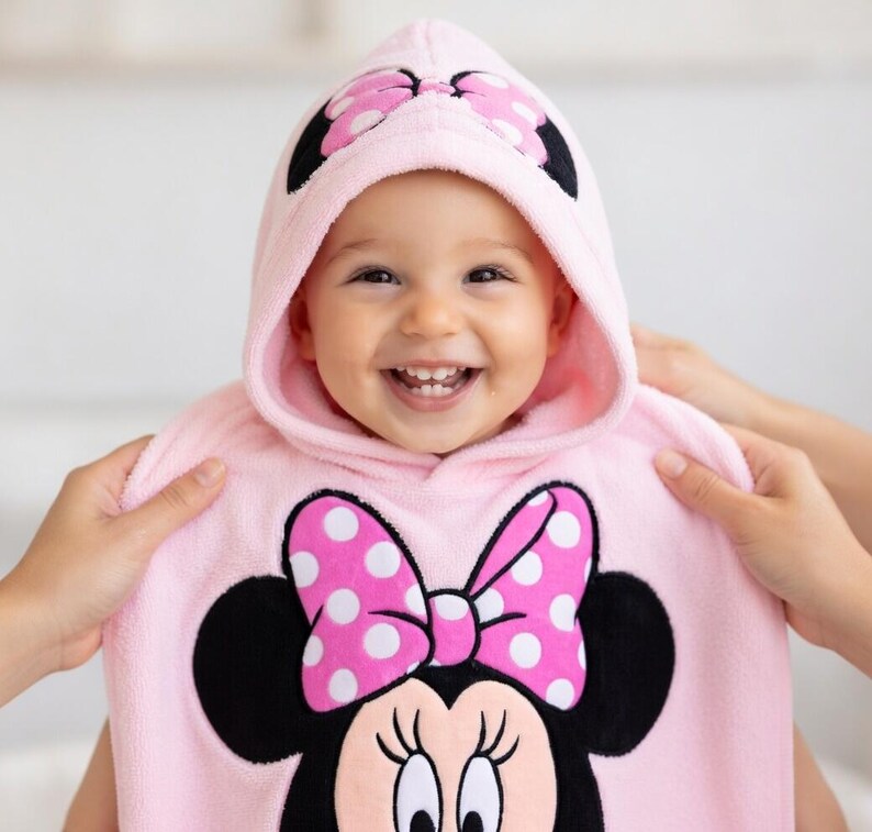 May include: A smiling baby wearing a pink hooded towel with a Minnie Mouse design. The towel features a large polka-dot bow and Minnie's face. The hood has a matching bow design. The baby is being held by two hands.