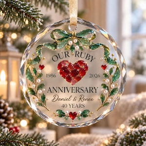 May include: A round, faceted glass ornament featuring a central red heart made of small stones. Text reads: OUR RUBY ANNIVERSARY, 1986-2026, Daniel & Renee, 40 YEARS. The design includes green painted leaves, white pearl-like accents, and a gold ribbon.