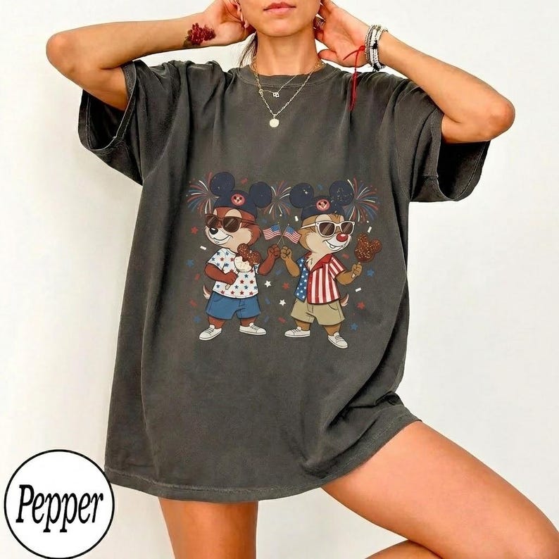 May include: Dark gray t-shirt featuring a graphic of two cartoon chipmunks wearing sunglasses and Mickey Mouse ears, holding American flags and treats. The chipmunks are dressed in patriotic outfits with fireworks in the background. The shirt has a relaxed fit.