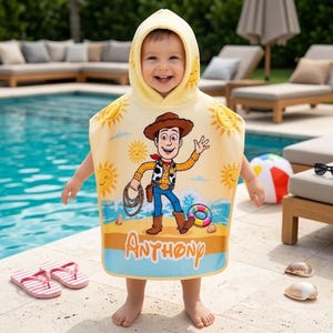 Puede incluir: Un ni&ntilde;o con una toalla de playa con capucha amarilla con un dise&ntilde;o de dibujos animados de Woody de Toy Story. La toalla presenta una escena de playa y el nombre "Anthony". Se ven chanclas de rayas rosas y blancas.