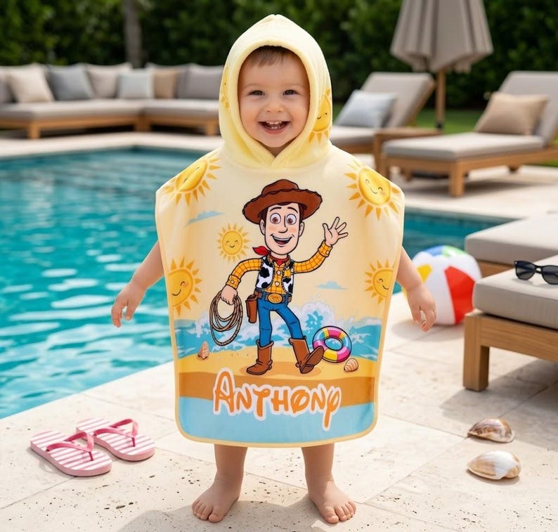Puede incluir: Un ni&ntilde;o con una toalla de playa con capucha amarilla con un dise&ntilde;o de dibujos animados de Woody de Toy Story. La toalla presenta una escena de playa y el nombre "Anthony". Se ven chanclas de rayas rosas y blancas.