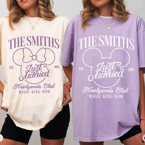 May include: Two oversized t-shirts, one ivory and one orchid, featuring a wedding-themed design. The design includes the text "THE SMITHS", "Just Married", and "Newlyweds Club", with a Mickey Mouse head silhouette. The shirts are for the "Magic King Dom" and the year "2025".