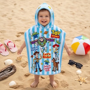 May include: A child wearing a blue and white striped hooded beach towel featuring Toy Story characters. The towel has images of Buzz Lightyear, Woody, and other characters. The child is standing on a sandy beach with flip-flops and a beach ball nearby.