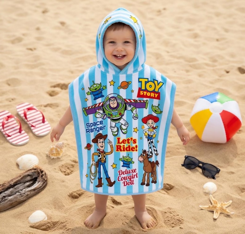 May include: A child wearing a blue and white striped hooded beach towel featuring Toy Story characters. The towel has images of Buzz Lightyear, Woody, and other characters. The child is standing on a sandy beach with flip-flops and a beach ball nearby.