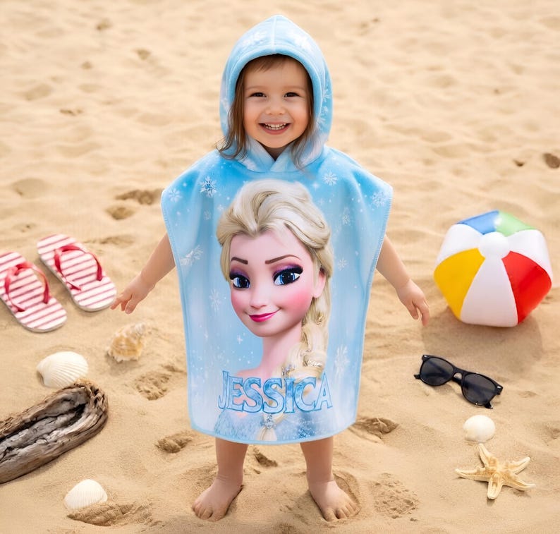 Puede incluir: Toalla de playa con capucha azul claro con un dise&ntilde;o de Elsa de Frozen y el nombre "JESSICA". Un ni&ntilde;o lleva la toalla en una playa de arena, con chanclas, un bal&oacute;n de playa y gafas de sol.
