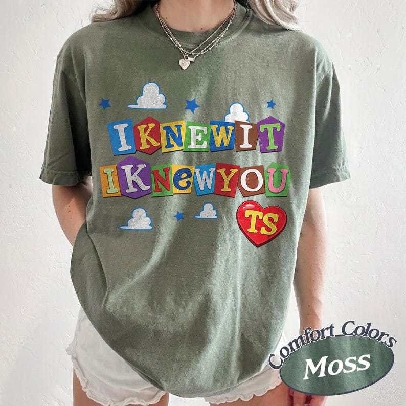 May include: An olive green short-sleeve t-shirt featuring a graphic print with the text "IKNEWIT IKNEWYOU" in colorful, block-lettered shapes. The design includes small white clouds, blue stars, and a red heart containing the yellow letters "TS".