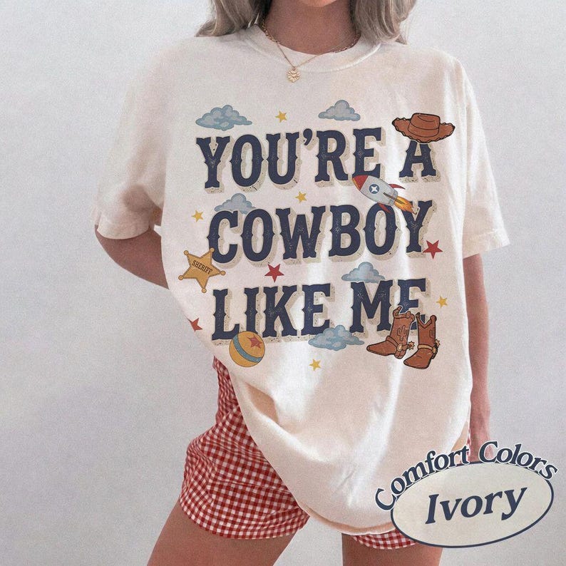 May include: An ivory short-sleeve t-shirt featuring the dark blue text "YOU'RE A COWBOY LIKE ME" surrounded by graphic illustrations of a brown cowboy hat, a rocket ship, a yellow sheriff star, a colorful ball, and brown cowboy boots with clouds.