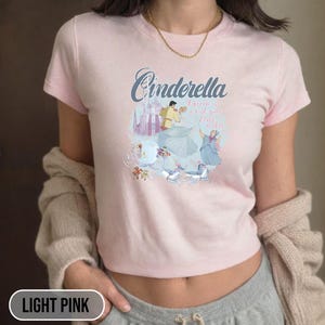 May include: Light pink t-shirt with a graphic of Cinderella and Prince Charming in a carriage with the text "A dream is a wish your heart makes".