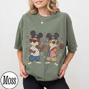 May include: Olive green t-shirt featuring a graphic of two cartoon chipmunks wearing sunglasses and Mickey Mouse ears, holding American flags and treats. The design includes fireworks and stars. The shirt has a round neckline and short sleeves.