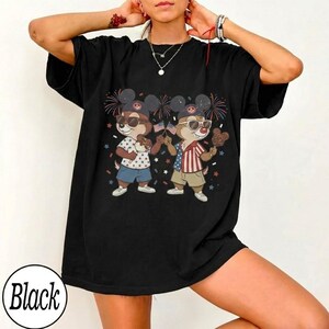 May include: Black t-shirt featuring a cartoon illustration of two chipmunks wearing sunglasses and patriotic outfits, holding ice cream and American flags. The chipmunks are set against a backdrop of fireworks and confetti.
