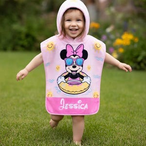 May include: A pink hooded beach towel featuring a Minnie Mouse graphic wearing sunglasses and a floatie. The towel has the name "Jessica" printed on the bottom. The background shows green grass and yellow flowers.
