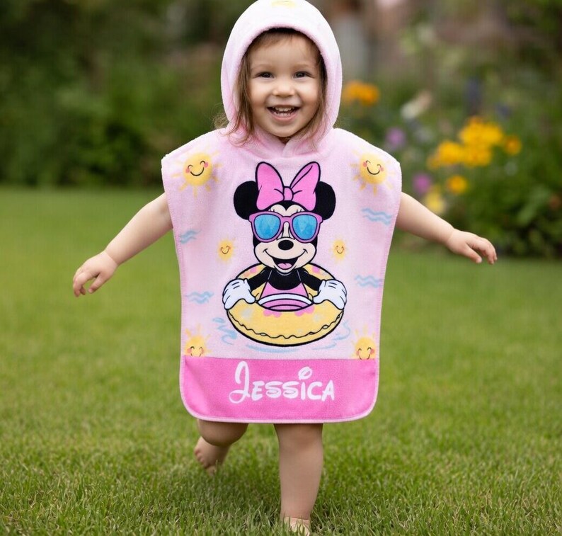 May include: A pink hooded beach towel featuring a Minnie Mouse graphic wearing sunglasses and a floatie. The towel has the name "Jessica" printed on the bottom. The background shows green grass and yellow flowers.