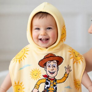Puede incluir: Un beb&eacute; sonriente con una toalla con capucha amarilla con una imagen de dibujos animados de Woody de Toy Story. La toalla presenta gr&aacute;ficos de sol y un pa&ntilde;uelo rojo. El beb&eacute; est&aacute; siendo sostenido por un adulto.