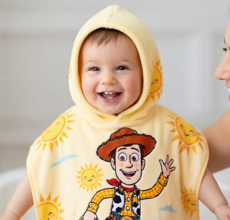 Puede incluir: Un beb&eacute; sonriente con una toalla con capucha amarilla con una imagen de dibujos animados de Woody de Toy Story. La toalla presenta gr&aacute;ficos de sol y un pa&ntilde;uelo rojo. El beb&eacute; est&aacute; siendo sostenido por un adulto.