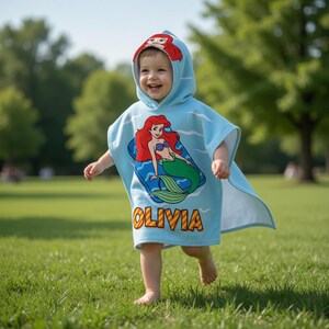 K&ouml;nnte beinhalten: Hellblaues Kapuzenhandtuch mit einem Cartoon-Meerjungfrau-Design mit roten Haaren und einem gr&uuml;nen Schwanz. Auf dem Handtuch ist der Name "OLIVIA" in Gelb aufgedruckt. Die Kapuze hat ein passendes Meerjungfrau-Design.